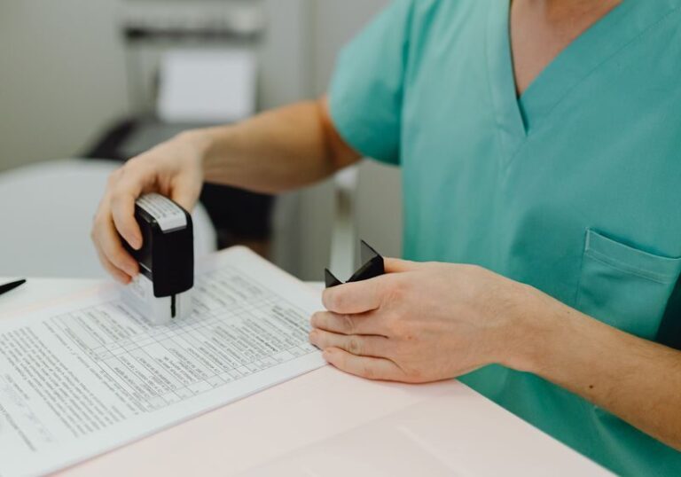 Healthcare professional wearing scrubs and mask reviewing documents at a desk indoors.